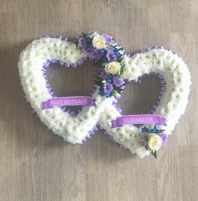 Double lilac heart - A beautiful and touching double heart tribute, symbolising everlasting love and an unbreakable bond. Crafted with a mass of pristine white chrysanthemums and delicately edged with soft lilac ribbon, this design is both elegant and deeply meaningful.

The two intertwined hearts are adorned with gentle accents of ivory roses, lilac blooms, and touches of soft blue florals, creating a calming and comforting colour palette. Finished with personalised ribbon banners, this tribute offers a truly personal way to honour a loved one and keep their memory close.

Perfect for funerals, memorials, or celebrations of life, this arrangement conveys love, peace, and remembrance with grace.

Customisation available – name ribbons, colour accents, and floral choices can be tailored to your wishes.