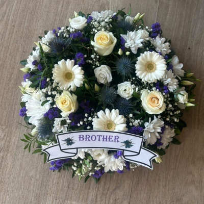 Scottish theme wreath - A beautifully crafted funeral tribute designed to honour the memory of a beloved brother. This classic arrangement features a harmonious mix of white gerberas, roses and chrysanthemums, complemented by blue eryngium, soft purple statice and delicate gypsophila. The combination of white and blue tones symbolises peace, remembrance and everlasting love.

Finished with fresh foliage and a personalised ribbon bearing the word “Brother”, this tribute offers a timeless and dignified farewell suitable for funerals, memorial services or celebrations of life.
Ribbon can be changed to your choice of name